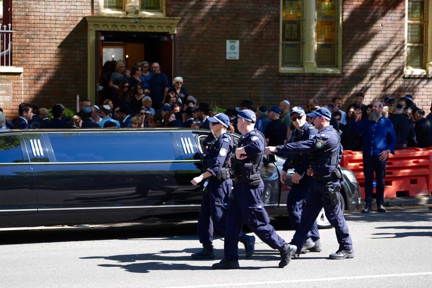 Cuatro agentes de policía pasan junto a un coche fúnebre negro frente a un edificio.