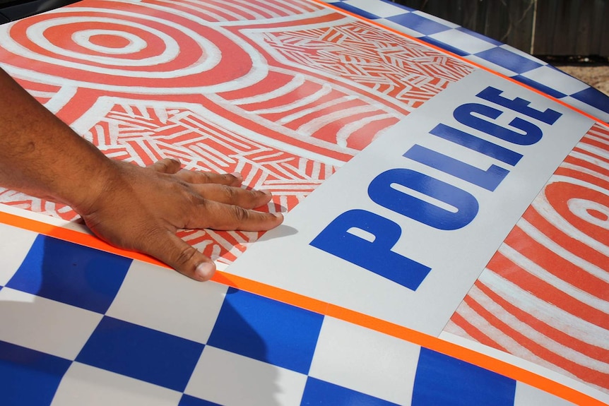 An Indigenous man's hand on the bonnet of  a police car painted with Aboriginal designs.