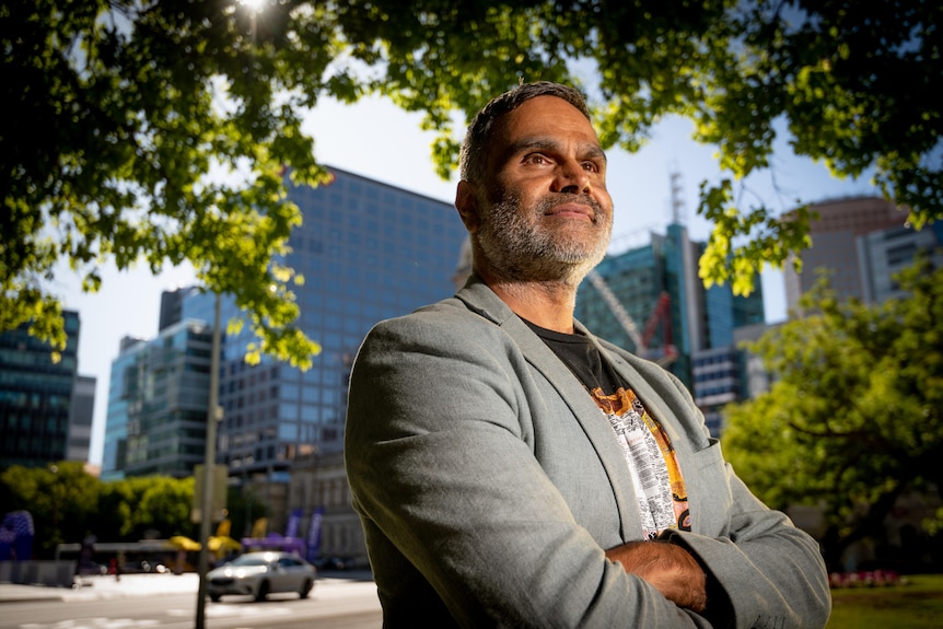A young Aboriginal man in a grey suit stands under a tree in front of some corporate buildings.