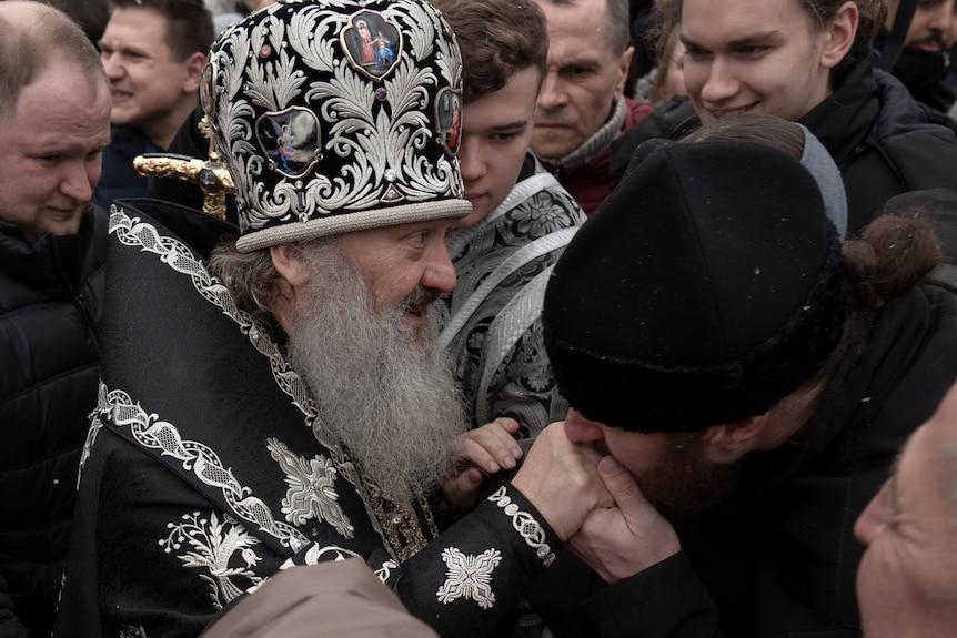 A priest with long grey beard and elaborate black and silver robes and hat is killed on the hand by a man in a black beanie.
