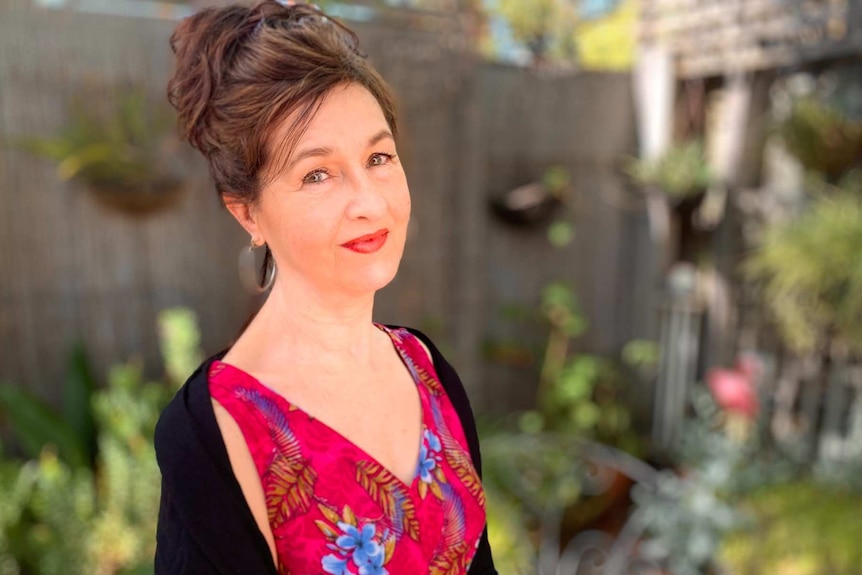 A woman stands in a garden and looks into the camera with a serious expression on her face.