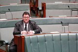 George Christensen sitting alone in the House of Representatives, looking into space as he holds his phone.
