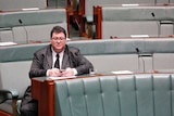 George Christensen sitting alone in the House of Representatives, looking into space as he holds his phone.
