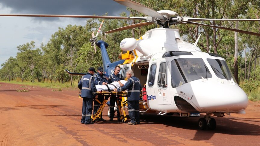 Large helicopter with four paramedics attending a patient on the gurney, on a red dirt road next to eucalypt trees