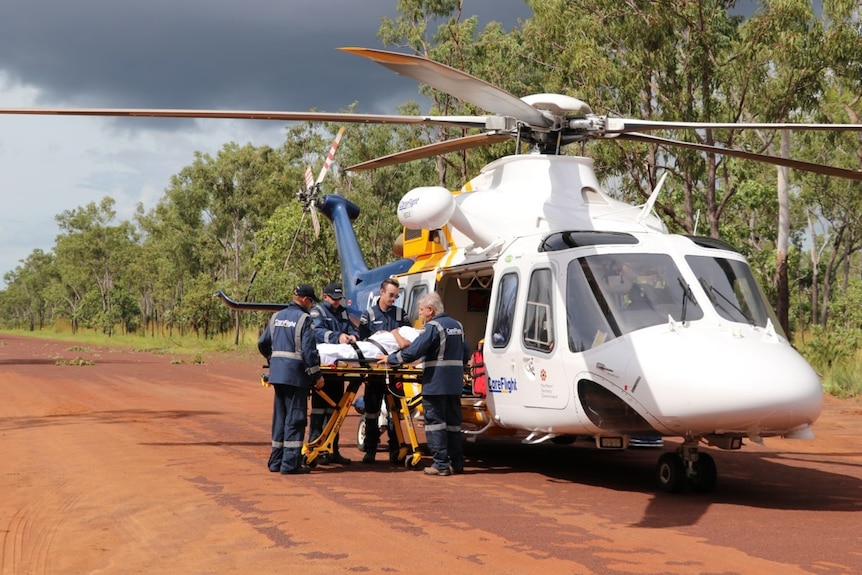 Large helicopter with four paramedics attending a patient on the gurney, on a red dirt road next to eucalypt trees
