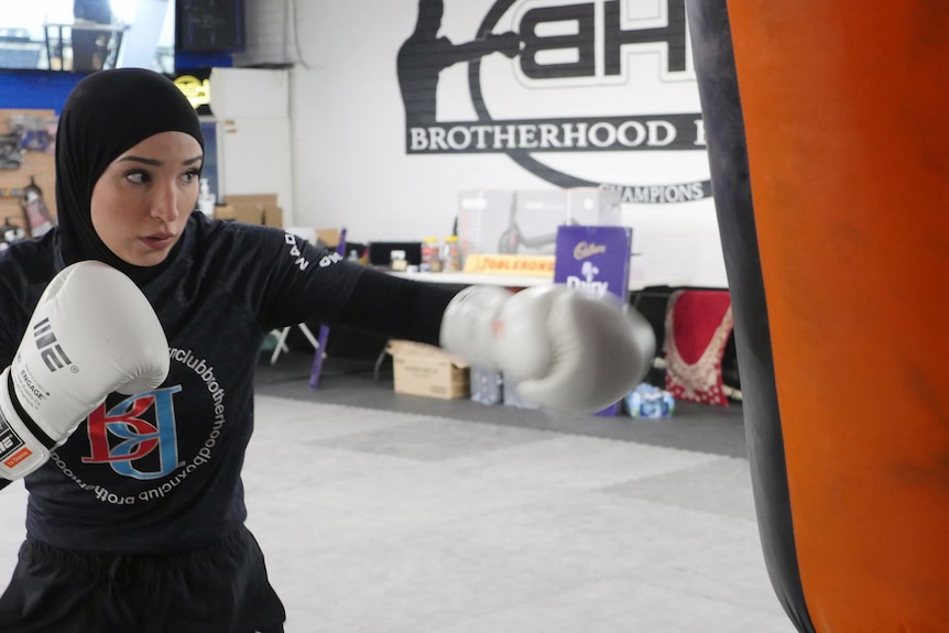 A female boxer wearing a headscarf and gloves hits a punching bag.