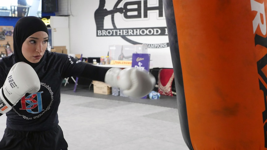 A female boxer wearing a headscarf and gloves hits a punching bag.