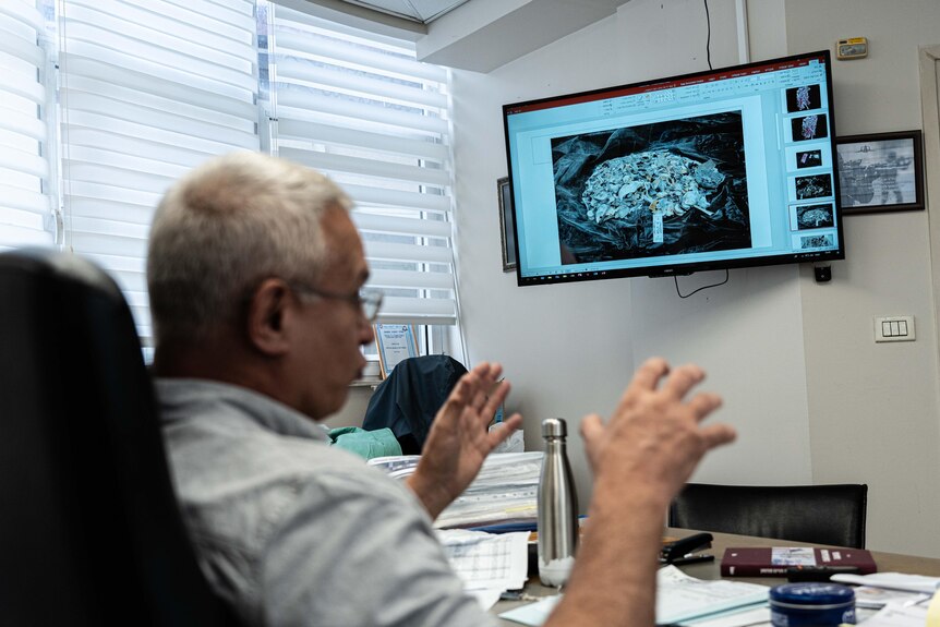 A man with white hair sits at his desk infront of a television screen displaying human remains.