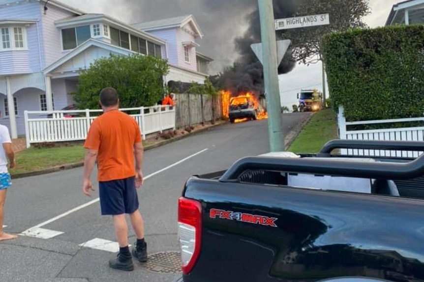 A man and a woman look on as a car burns down the street. 