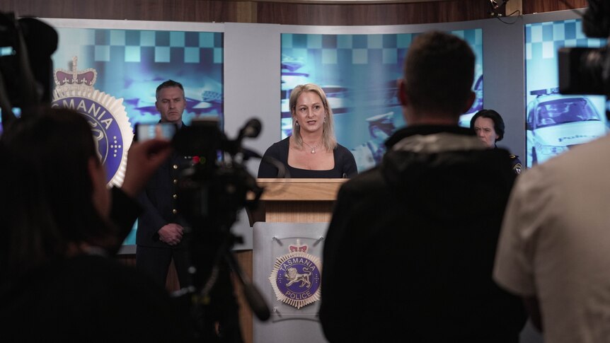 Blonde woman standing at podium and speaking with police officers behind her.