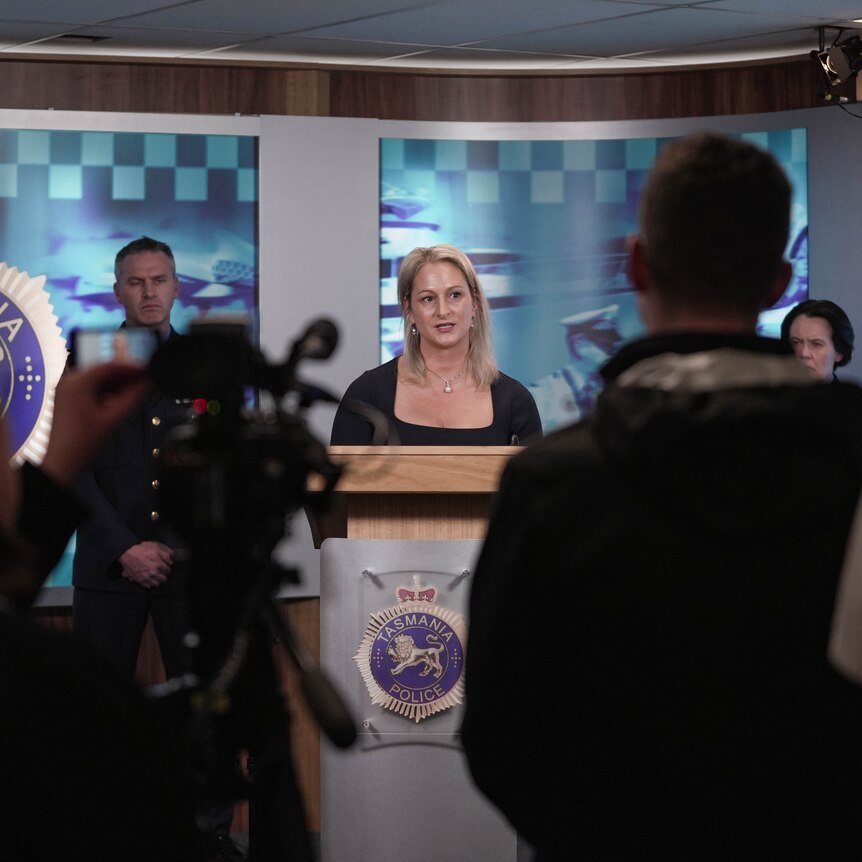 Blonde woman standing at podium and speaking with police officers behind her.