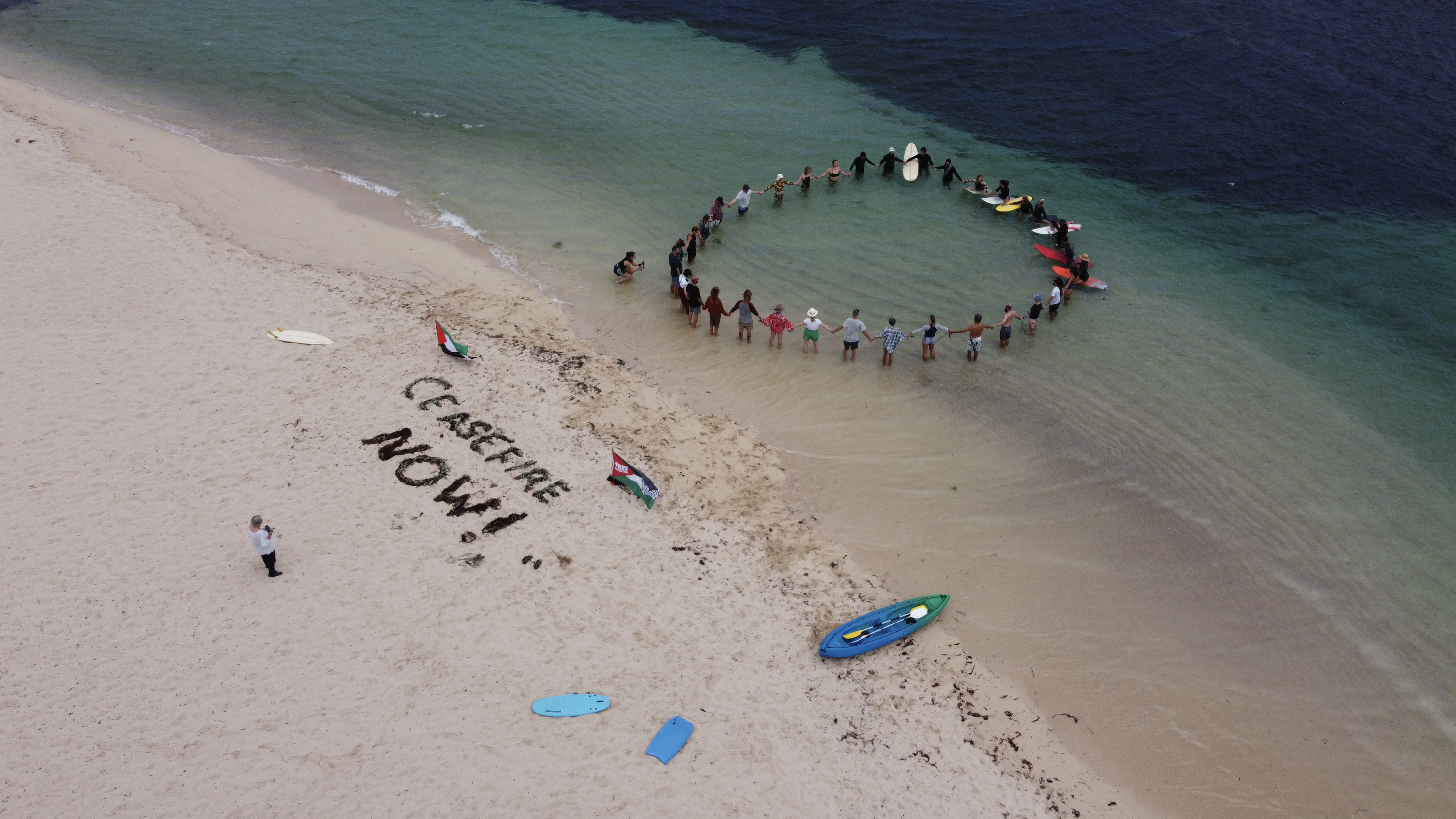 About 30 people form a circle in the water on surfboards or standing. On the beach, 'Ceasefire Now' has been spelt in seaweed.