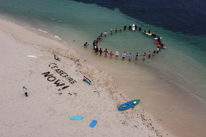 About 30 people form a circle in the water on surfboards or standing. On the beach, 'Ceasefire Now' has been spelt in seaweed.