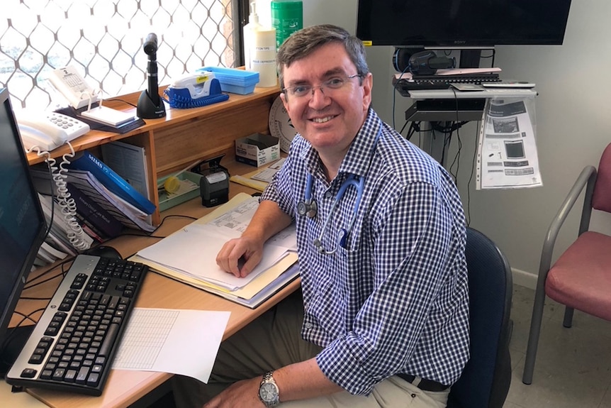 Dr Adam Coltzau wearing a blue chequered shirt, a stethoscope around his neck sitting in his consulting room.