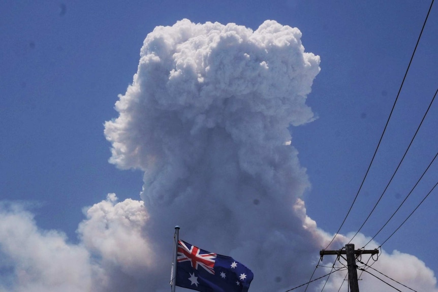 An Australian flag flies high on a flag pole near a powerline with a large plume of dark smoke in the background.