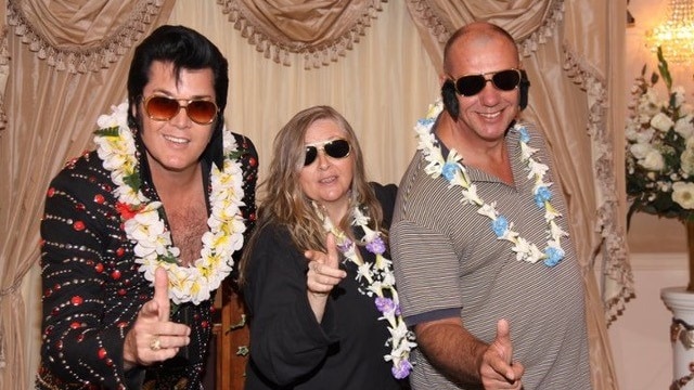 An older couple and a man dressed as Elvis, all wearing sunglasses, lean towards the camera