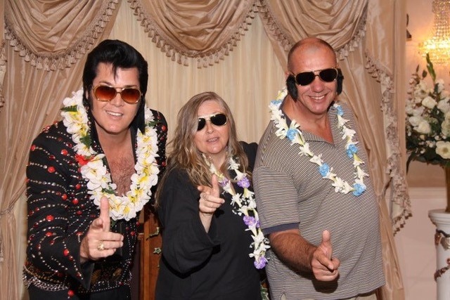 An older couple and a man dressed as Elvis, all wearing sunglasses, lean towards the camera