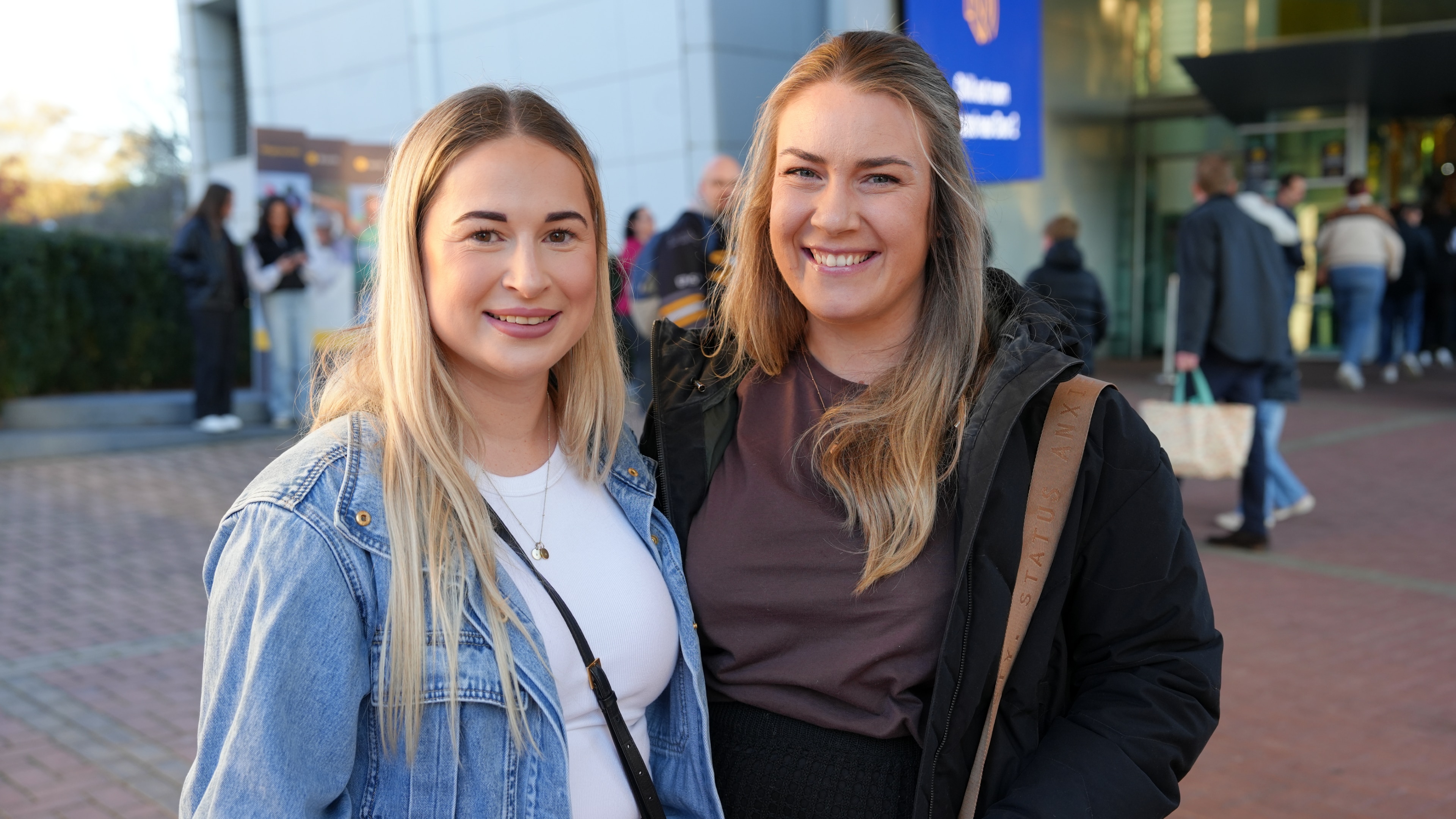 Two people with long blonde hair stand outside smiling, near the stadium.