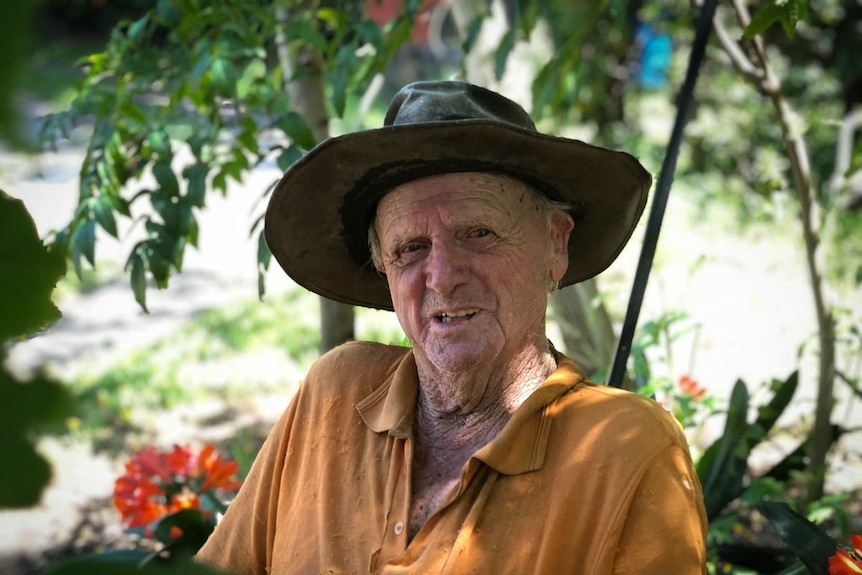 An elderly man with a hat sits on a chair surrounded by flowers