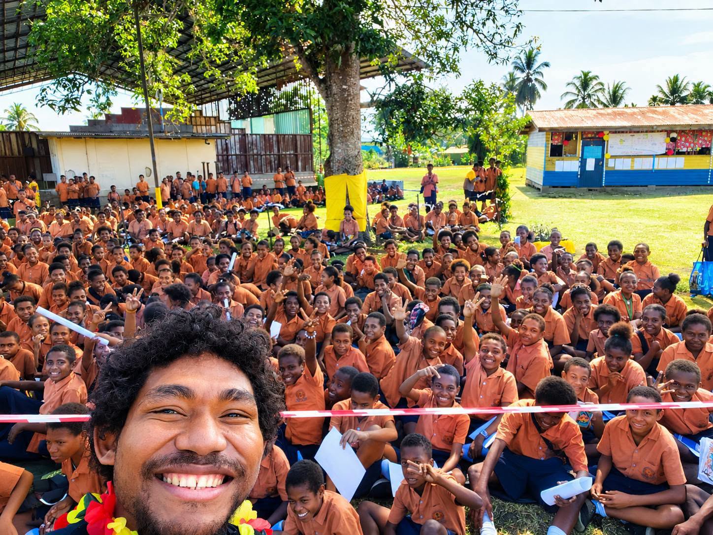 A Papua New Guinean man takes a selfie with his face in the forefront and a group of children in brown uniform sitting on grass 