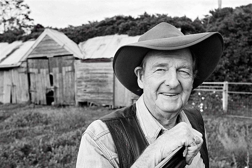 A black and white photo of an older man with a wide-brimmed hat standing in front of a ramshackle shed, smiling.