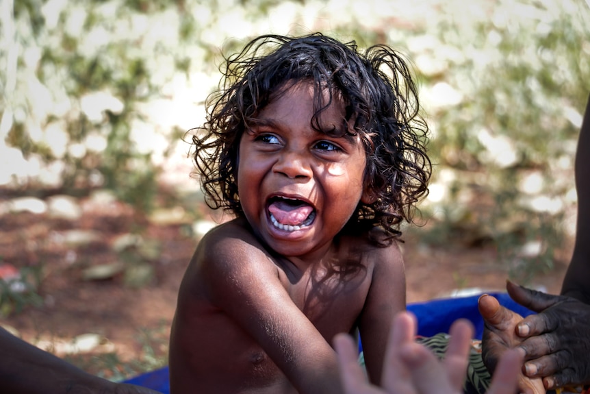 A happy boy smiles at the camera. He is not wearing a shirt and looks off camera.