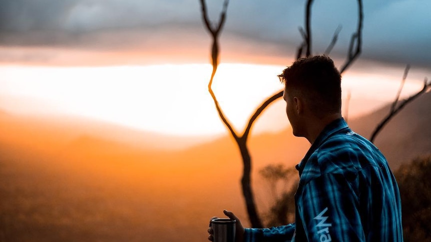 A man sits on a rock at sunrise, holding a travel mug.