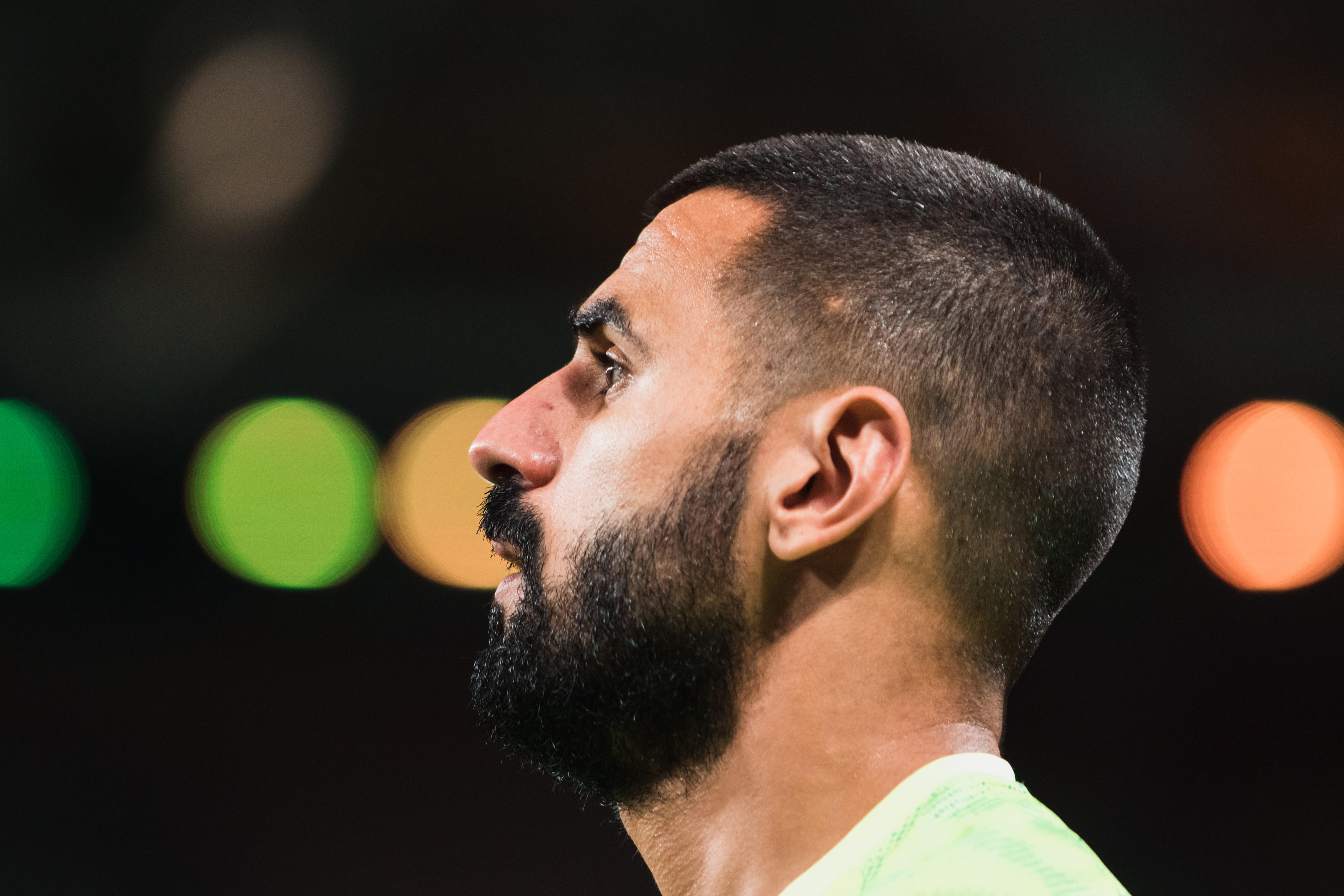 A man with a dark beard and wearing a bright green shirt looks out of frame with lights behind him