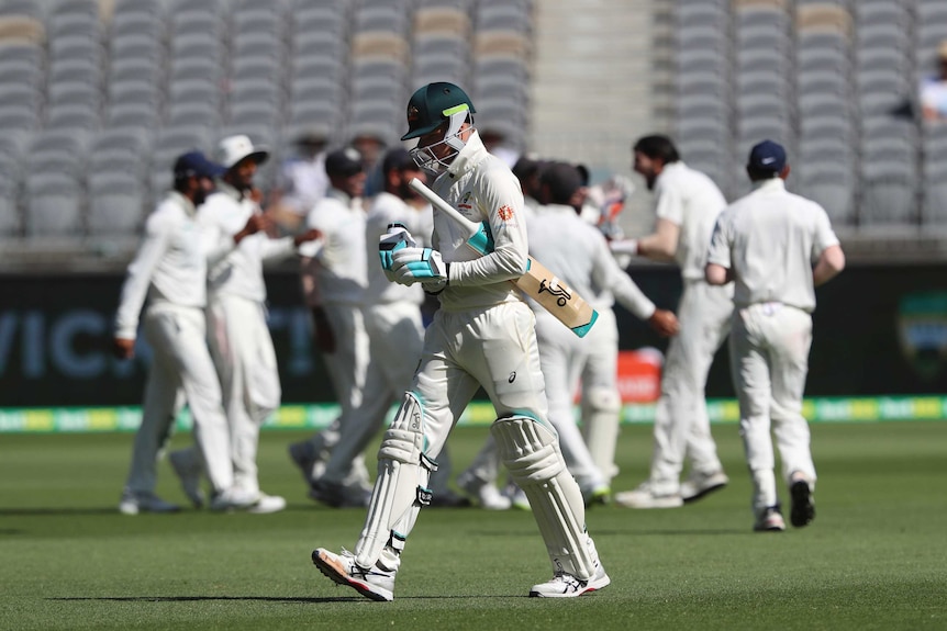 Australian batsman Peter Handscomb walks off the ground with his bat under his arm while Indian players celebrate.
