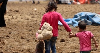 A older girl and younger boy hold teddies and hold hands as they walk with their backs to the camera.
