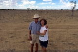 Husband and wife farmers standing in a dry paddock with brown dirt and short stubs of black grass.