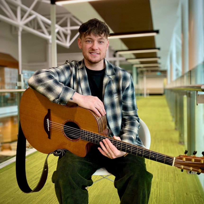 musician Ryan McMullan sits holding his guitar and smiles at the camera