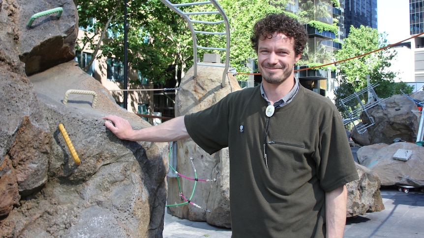 A man stands smiling next to a rock with metal handles sticking out