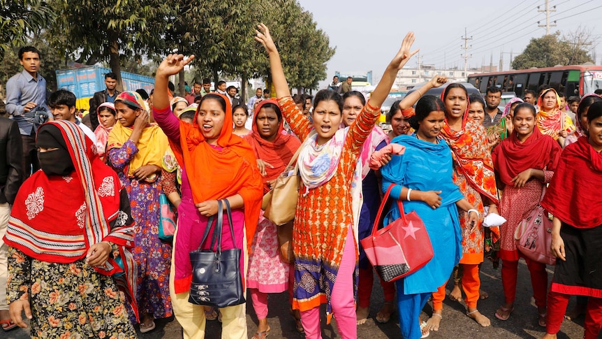 On a Bangladeshi street a large group of women in saris and veils lift their hands and shout in protest.