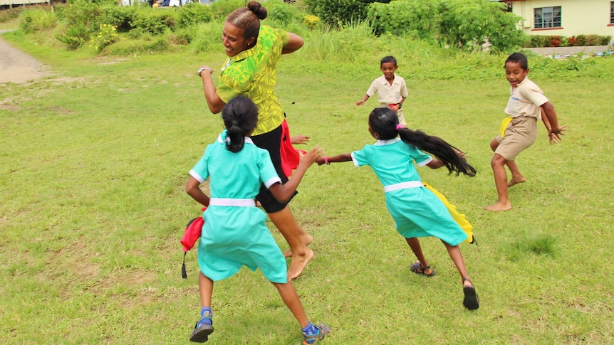 A woman with her hair in a bun, wearing a green floral shirt and dark skirt tries to escape girls in school uniform.