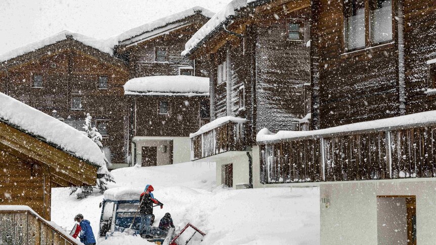 Wooden buildings are covered in snow as snow also falls in front of the camera lens
