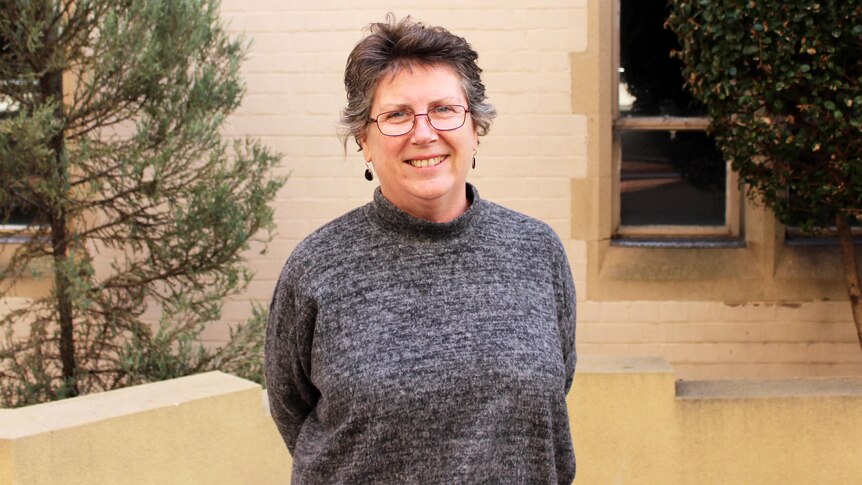A photo of a women with glasses and earrings wearing a grey sweater stands outside a sandstone building