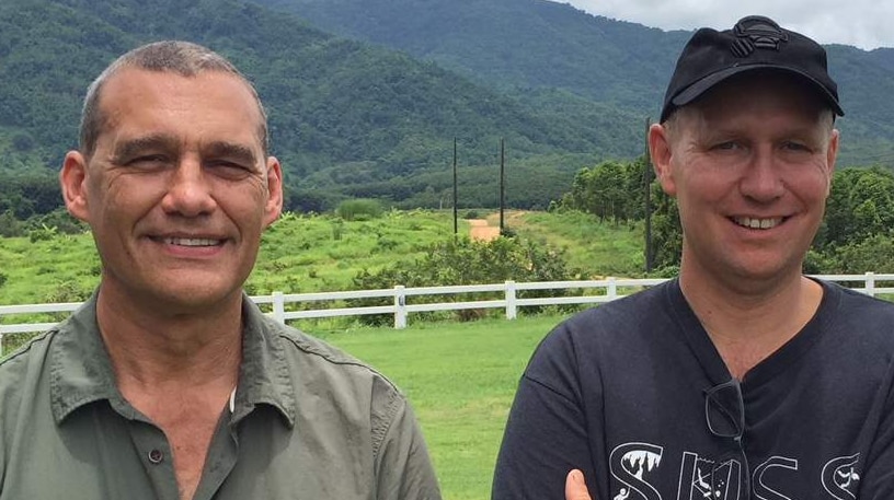 Two men stand in front of a lush green field at the bottom of a mountain in Thailand.