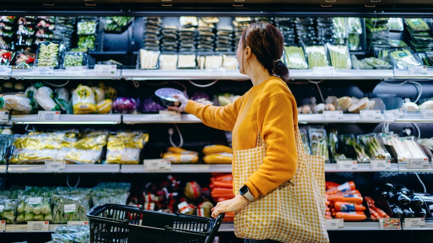 A woman in yellow jumper holding shopping bag faces a supermarket shelf full of colourful wrapped foods.