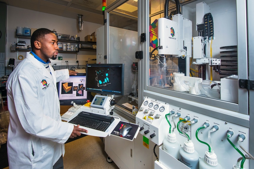 A young African American man in a lab coat works on a 3D printer in a laboratory.