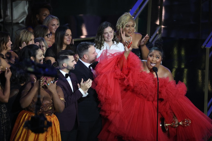 Lizzo wearing a red dress on stage, lots of people behind her