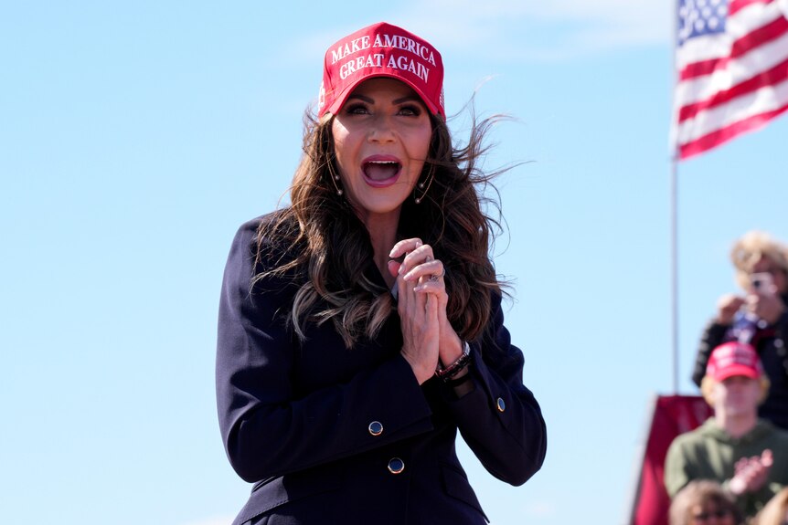 Kristi Noem stands outside, wearing a red 'Make America Great Again' baseball cap. 