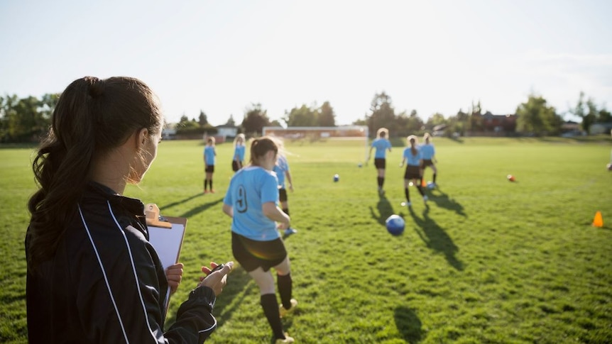 A female soccer coach with her back to the camera holding a stopwatch and watching teenage girls train.