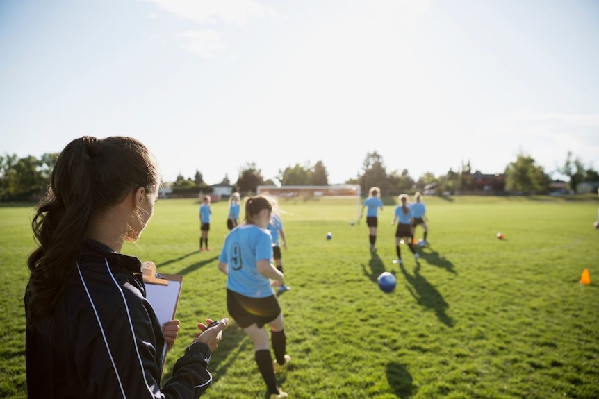 A female soccer coach with her back to the camera holding a stopwatch and watching teenage girls train.
