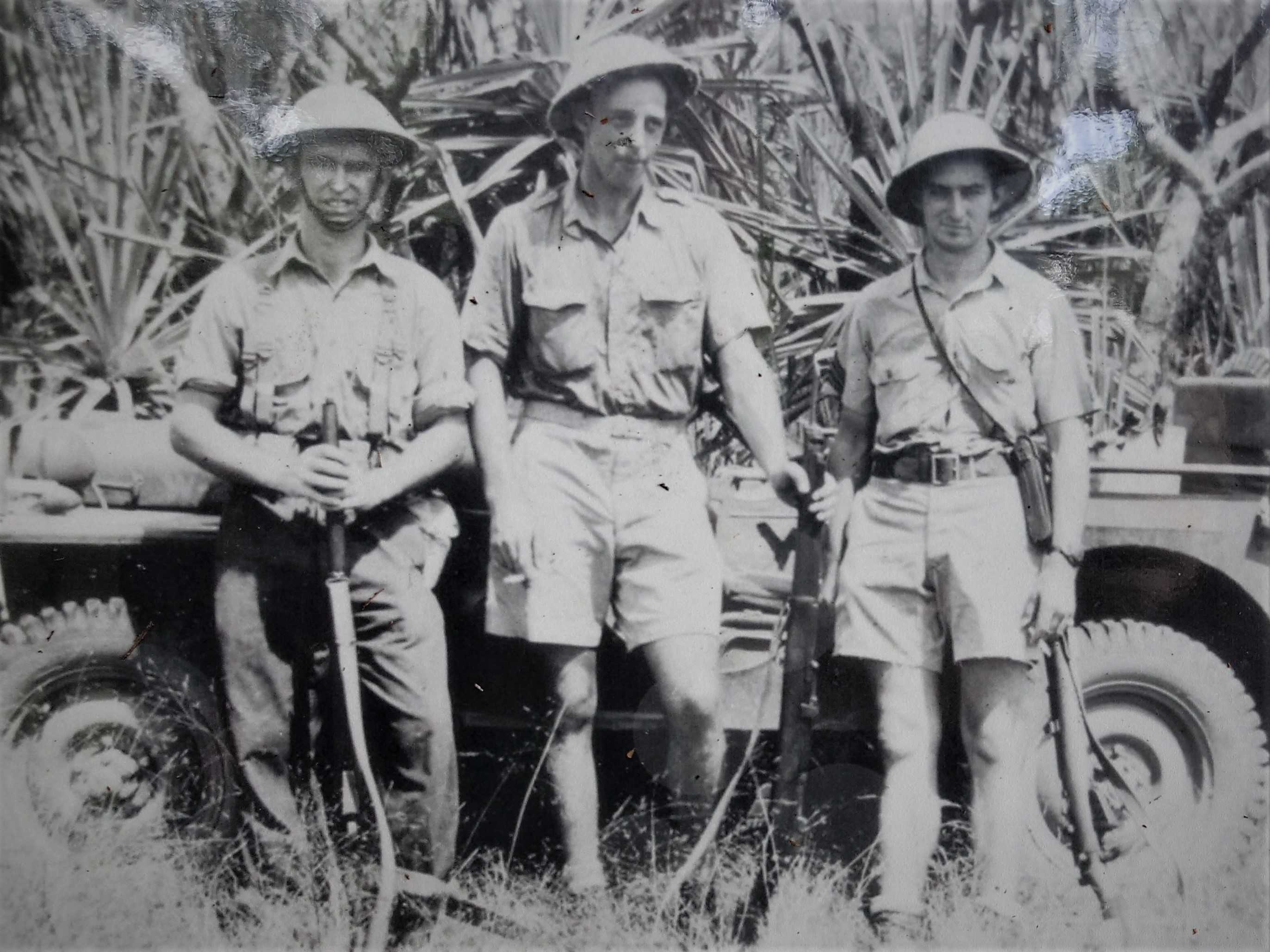 Monochrome of three American soldiers in uniform standing in the bush holding rifles with a vehicle behind them.