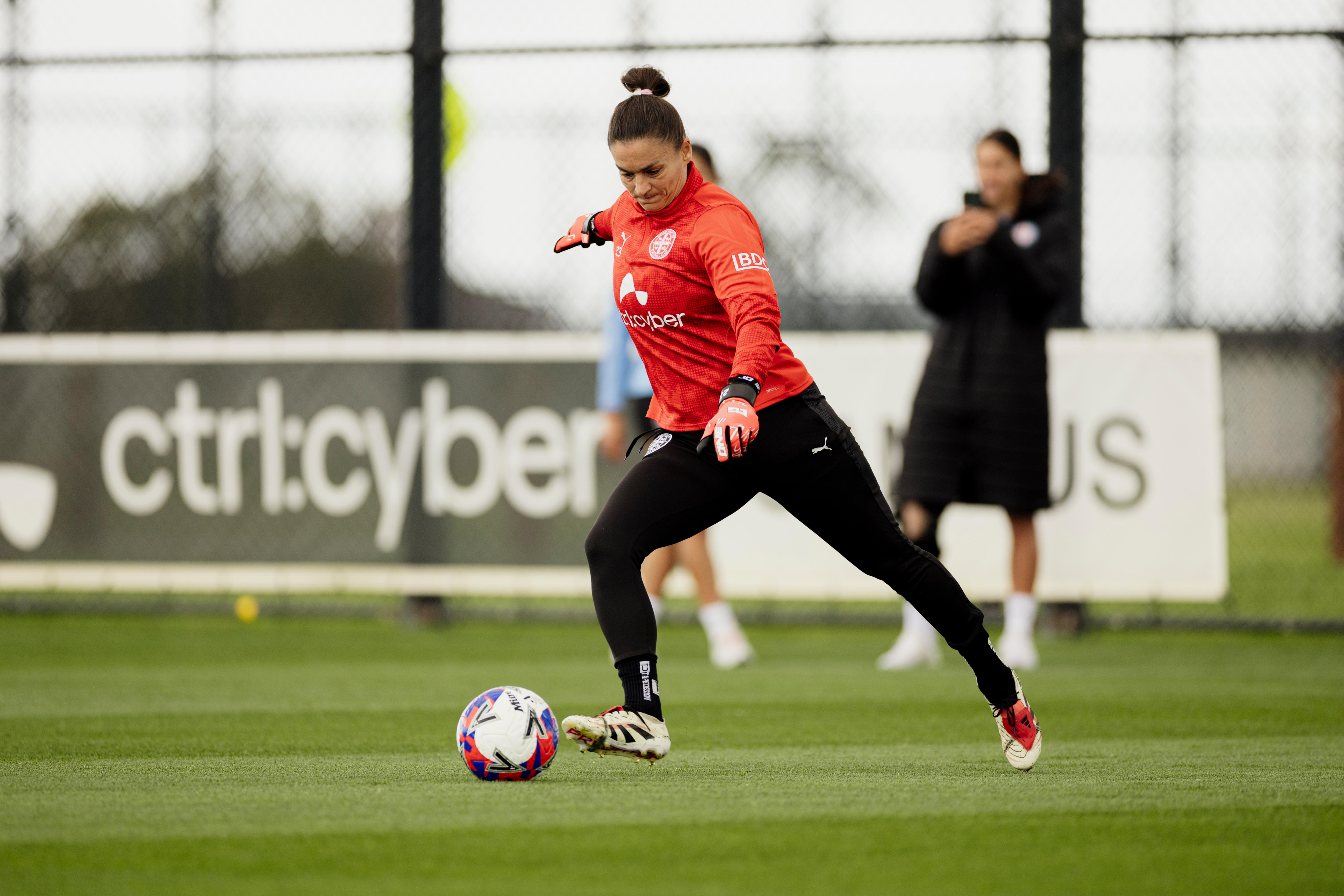 Ms Barbieri on a green soccer field in a red longsleeve and black leggings kicking a ball.