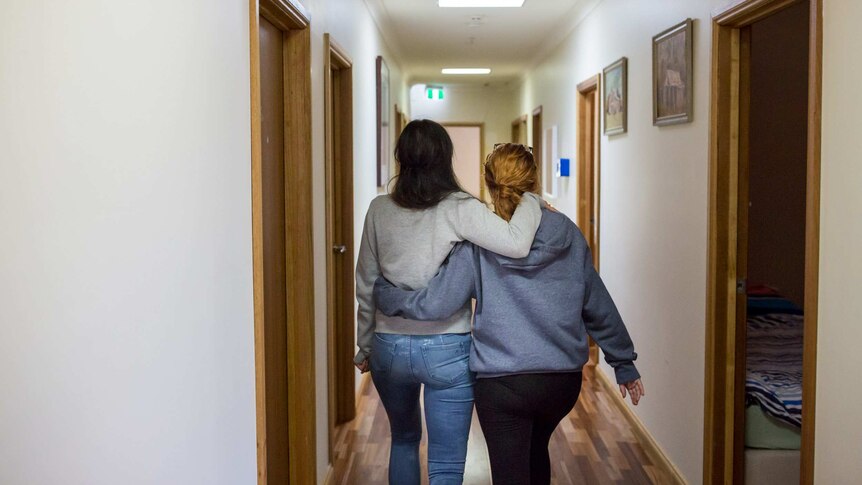 Two women walk down a hallway, holding one another.