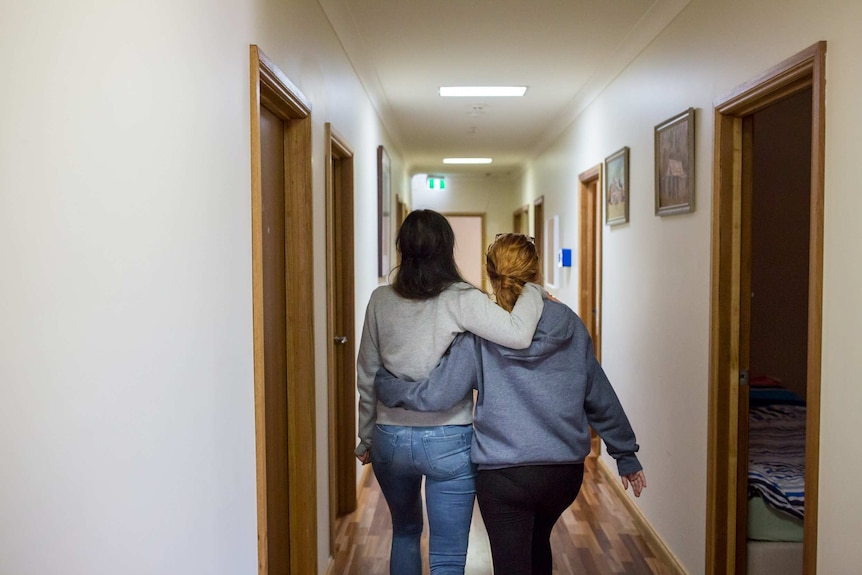 Two women walk down a hallway, holding one another.