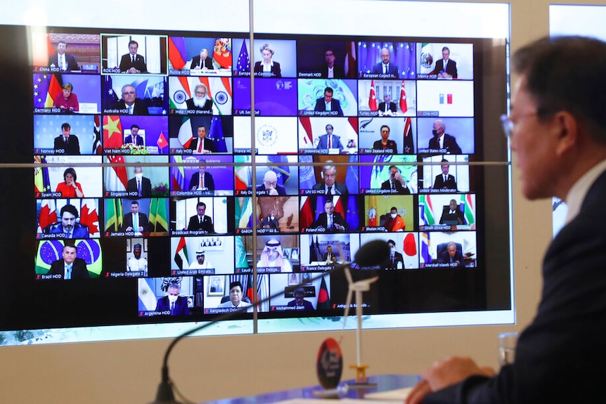 Mirando el hombro de Moon Jae-in, mira una gran pantalla de televisión que muestra filas de pequeños rectángulos que muestran a los líderes mundiales.