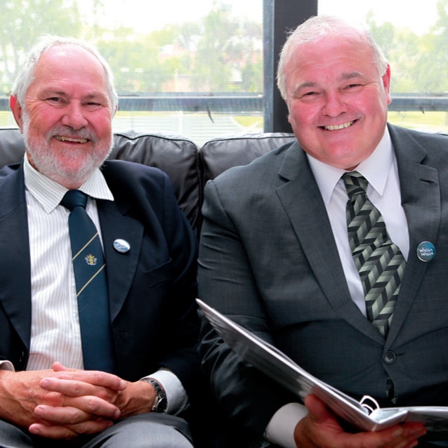 Two older men in suits, sitting next to each other and smiling.